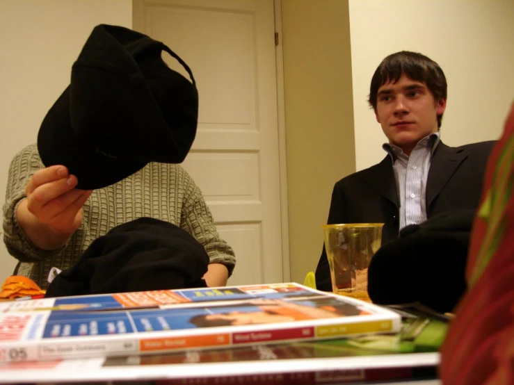 two men sitting around table with a stack of books and a hat on it