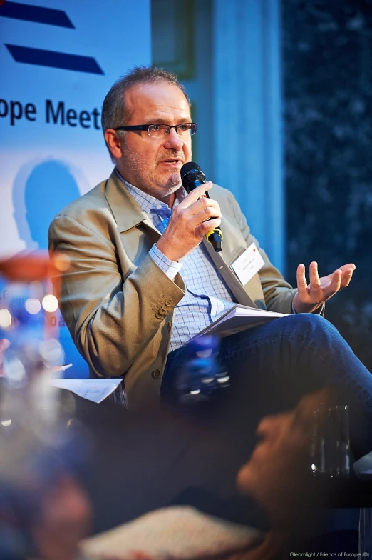 man holding a microphone speaks into his right hand while seated in front of a panel