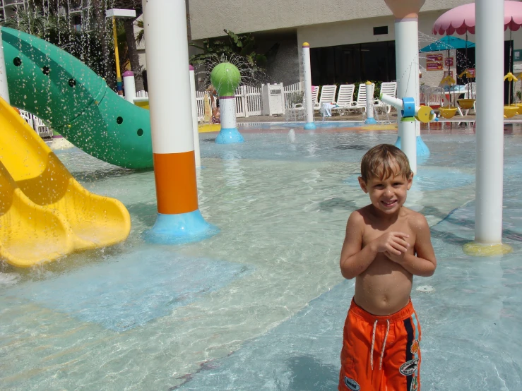 a boy in swim trunks standing near the edge of a swimming pool with a slide and splashpark in the background