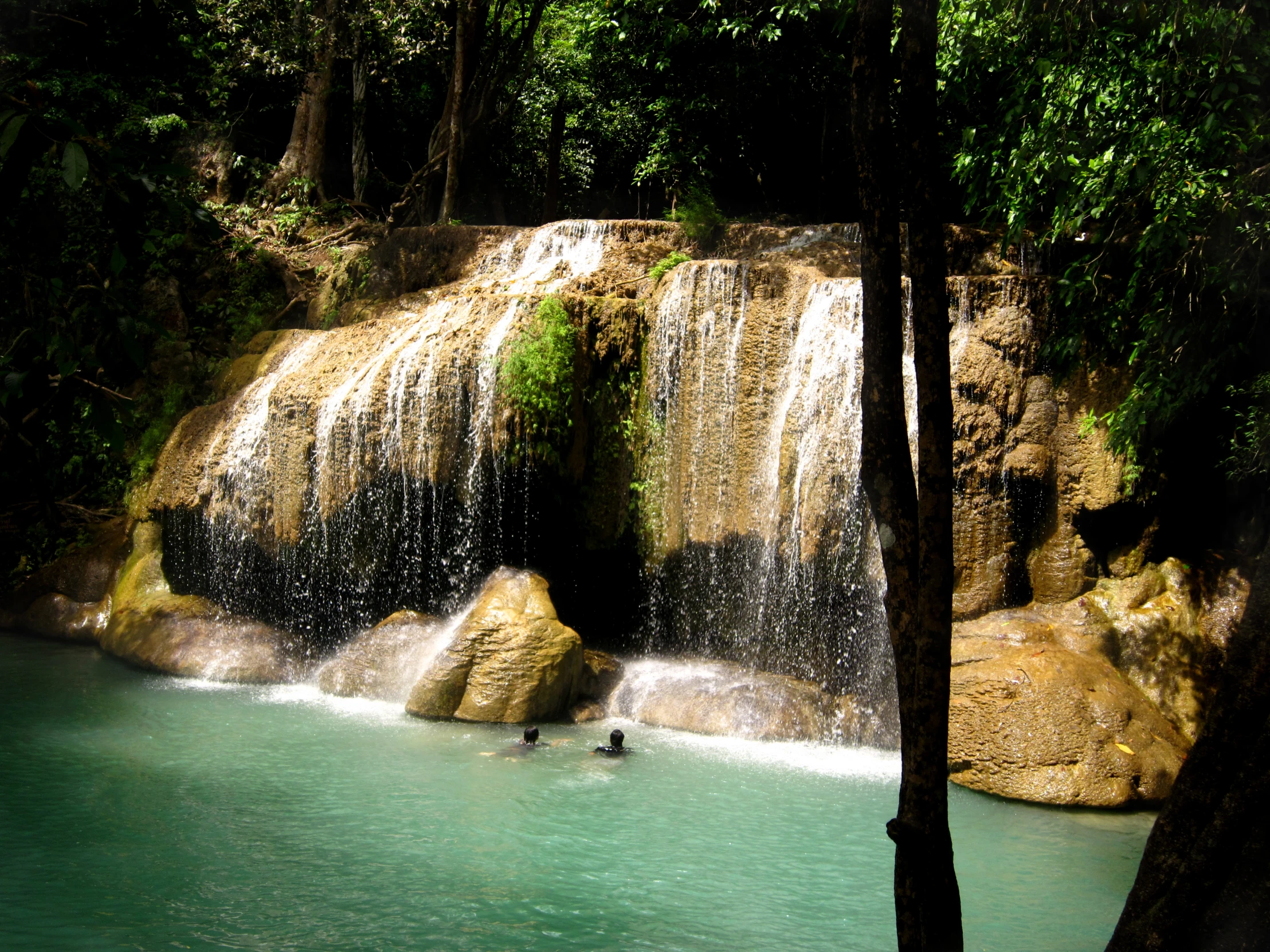 two people swim in a river next to the waterfall