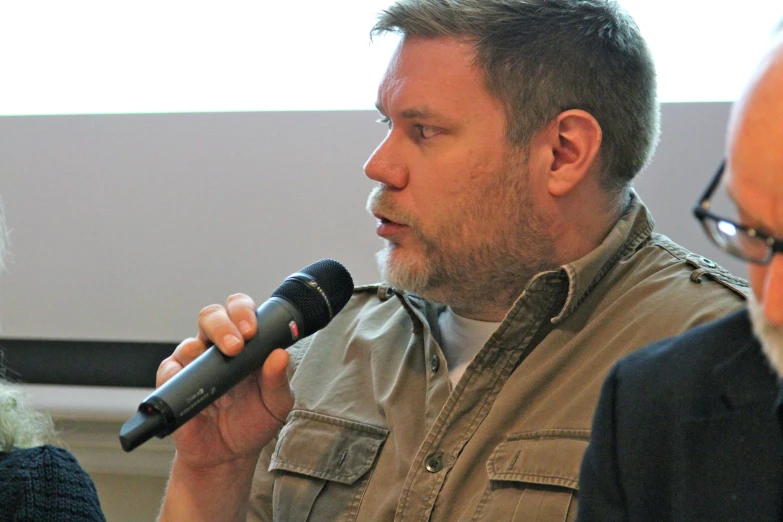 a man wearing a beige shirt and glasses speaks into a microphone