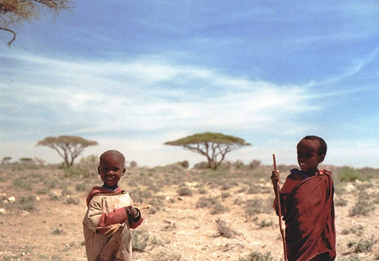 two boys stand on dirt with one of them holding a stick