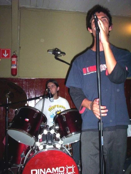 two people singing into some microphones while another man plays the drum