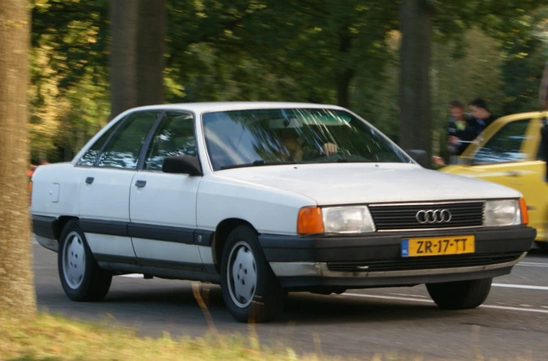 an old white car driving down a road by some trees