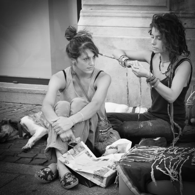 two women sitting on a sidewalk and one is iding the hair of another
