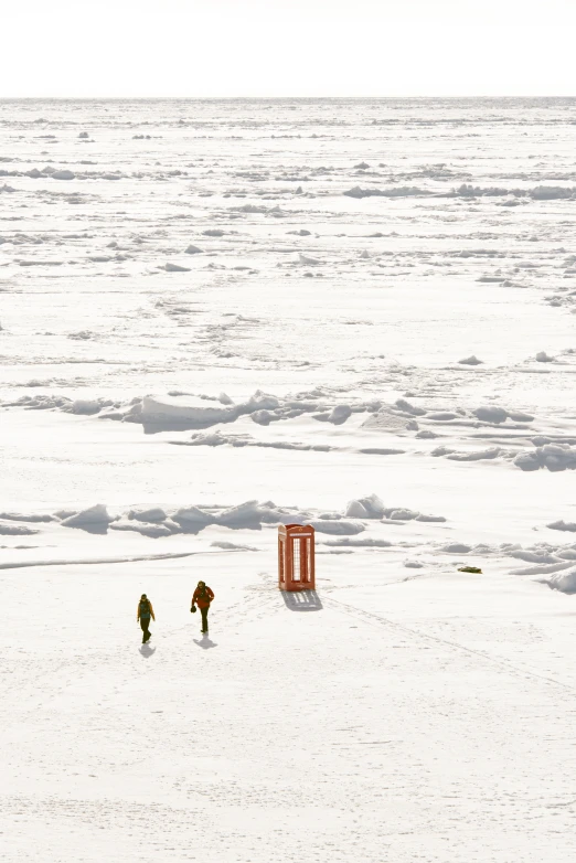 two people walking across snow covered ground next to a red phone booth