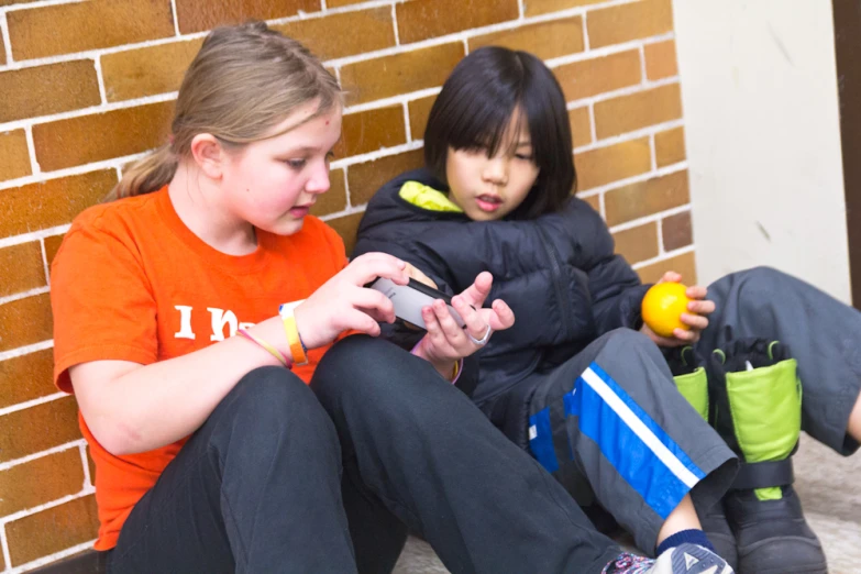 two little girls sitting against a brick wall on their cell phones