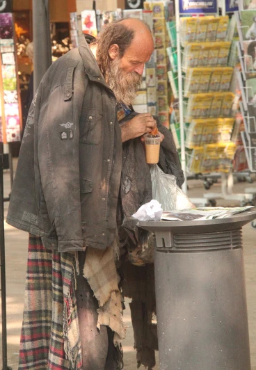 a man with a beard on a street corner, is picking up a garbage can