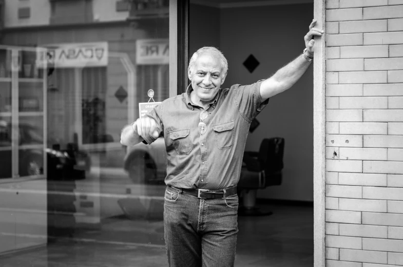 an old man in jeans and a on down shirt stands next to a wall holding a glass door