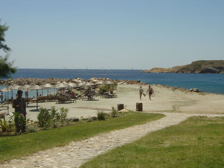 people sit on tables at the edge of a beach area with benches and water in the background