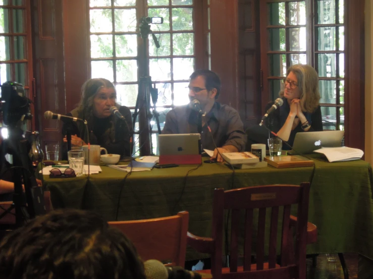 three people seated at a table while a person with a laptop talks on a microphone