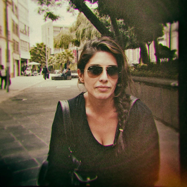 a woman wearing sunglasses standing near a tree on the sidewalk