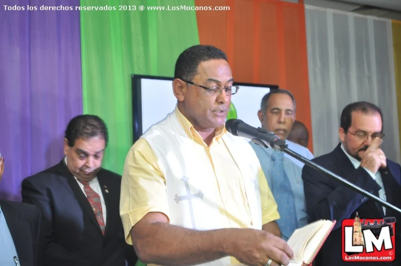 a man speaking into a microphone with men holding an open bible in front of him