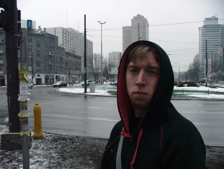 man in black jacket and red hood standing next to street with buildings and light pole