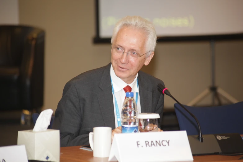 a man in suit and tie sitting at a table with a microphone and white board