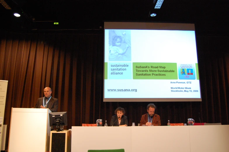 the panel at a conference are sitting next to a large screen