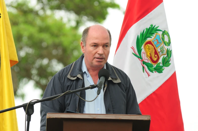 a man speaking at a podium with a flag and trees in the background