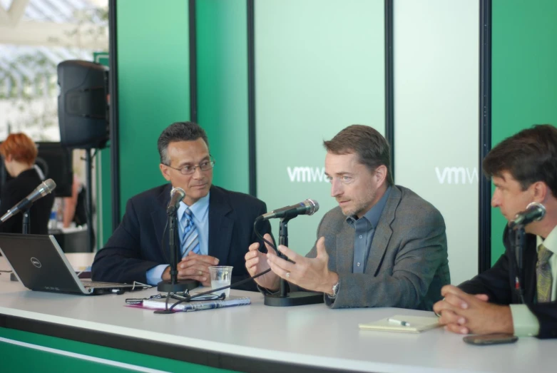 two men at a table with a laptop and microphone