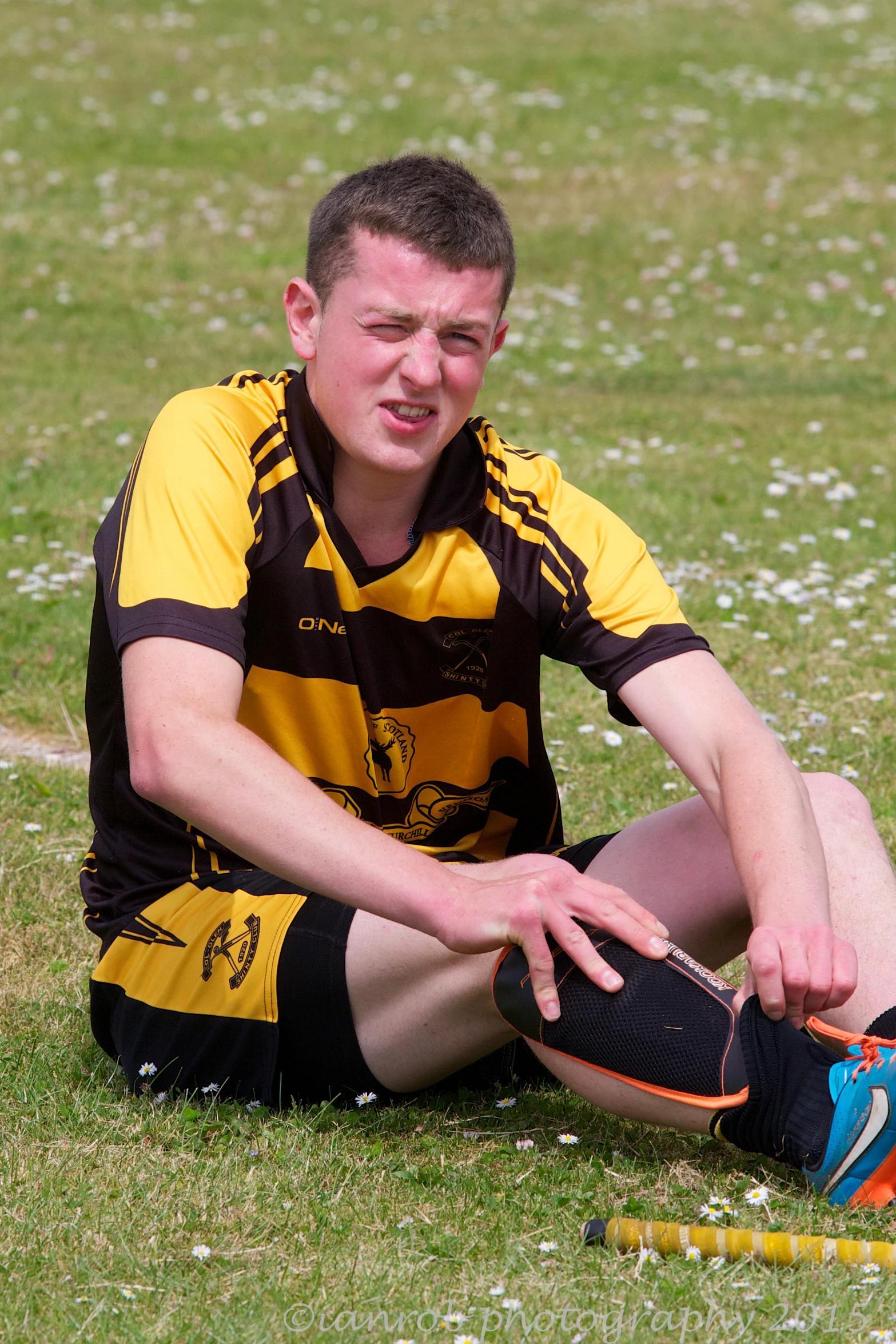 young man wearing black and yellow uniform sitting on green field