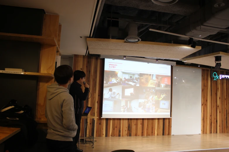 two people stand before a projector screen in front of a large room