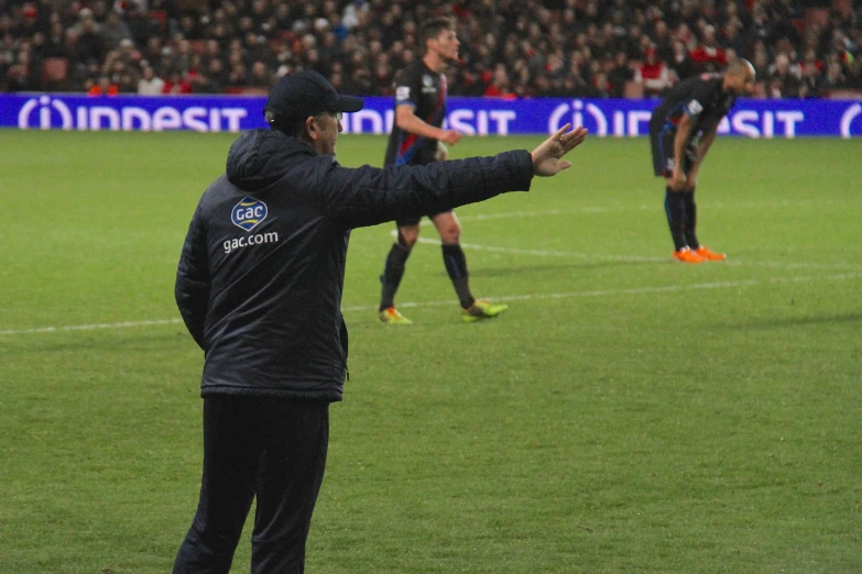 two men at a soccer field in black jackets with one holding his hand out