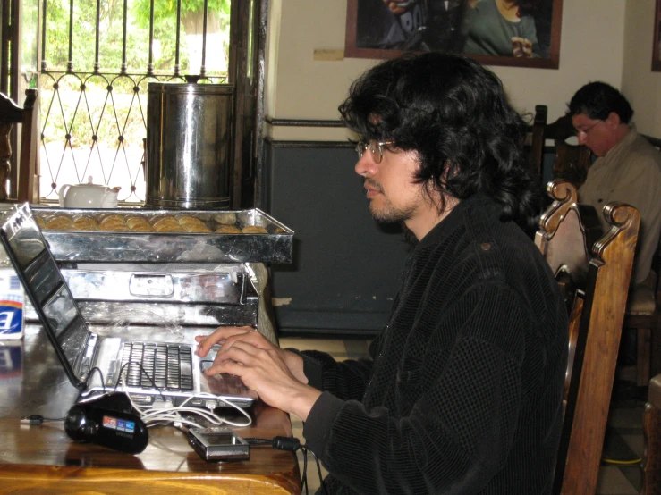a man with black hair sitting at a table working on a laptop computer