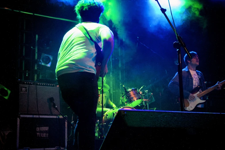 a man in yellow shirt playing guitar while another man in white shirt stands behind him on stage