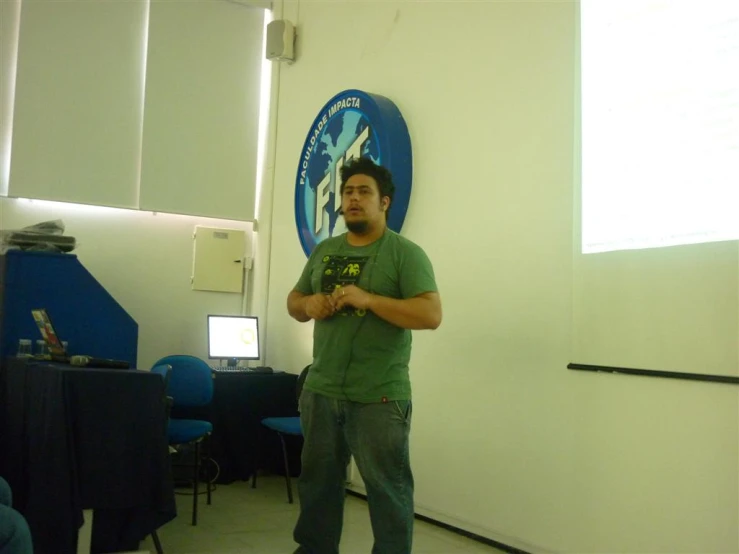 man with hands together looking to his left standing next to a room with projection screen