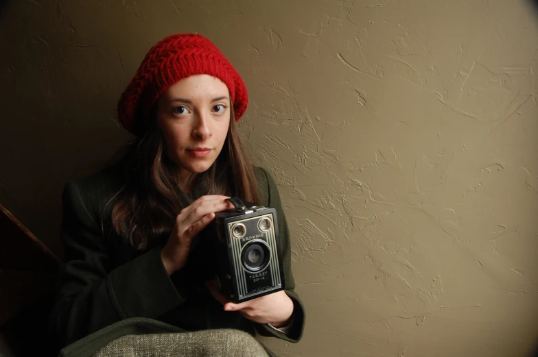 a girl in a red knit hat holds up an old - fashioned camera