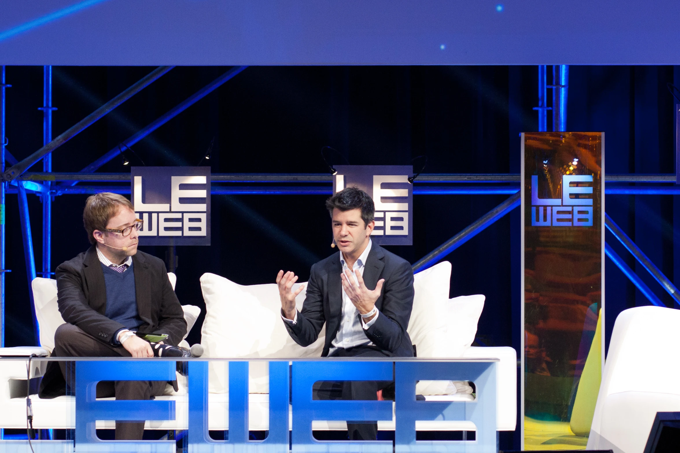 two men sitting on couches in front of blue and white speakers