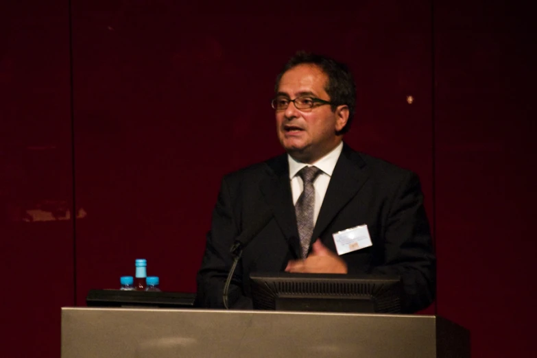 man wearing a black suit and tie standing at a podium