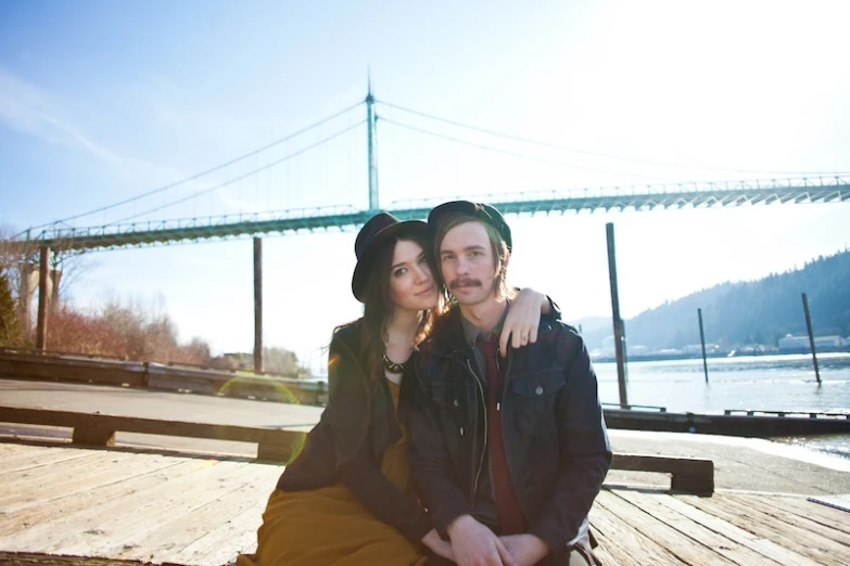 a man and woman in a black jacket sitting on a bench by the water