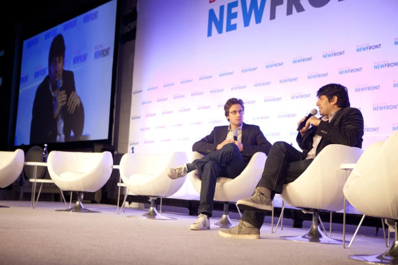 two men are sitting on chairs speaking to a crowd