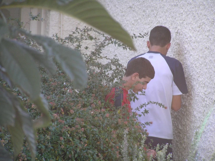 two boys stand together against a stucco wall, one is looking down and the other one is leaning against a plant