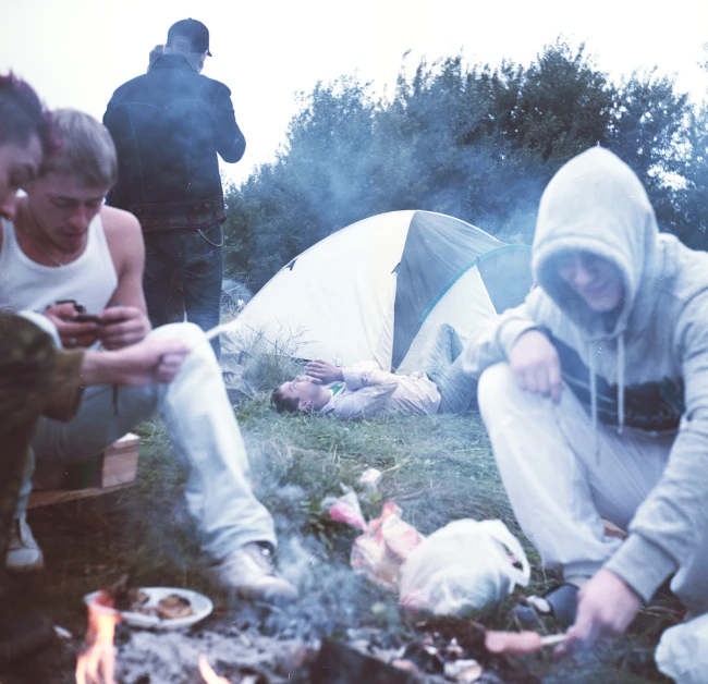 a man in hoody sweatshirts cooking food while others sit next to tent