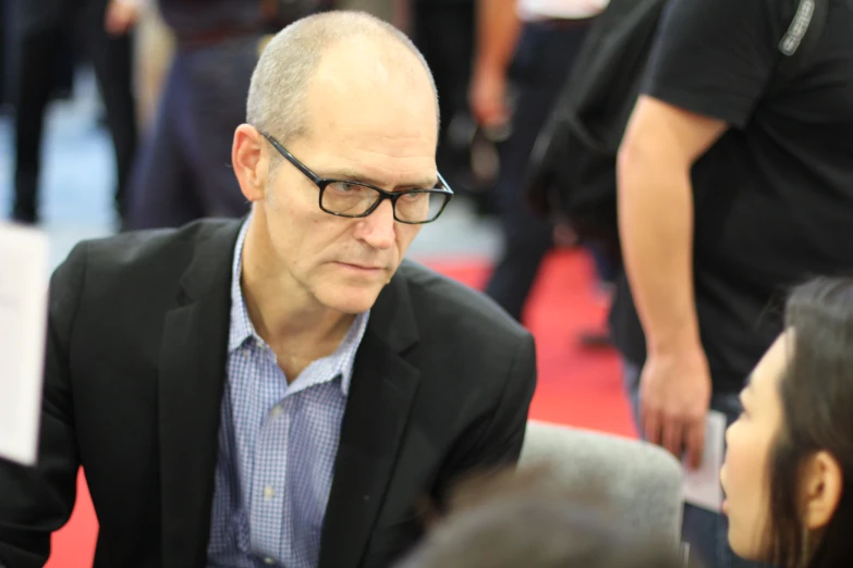 the man wearing glasses sits on a red carpet in a crowd of people