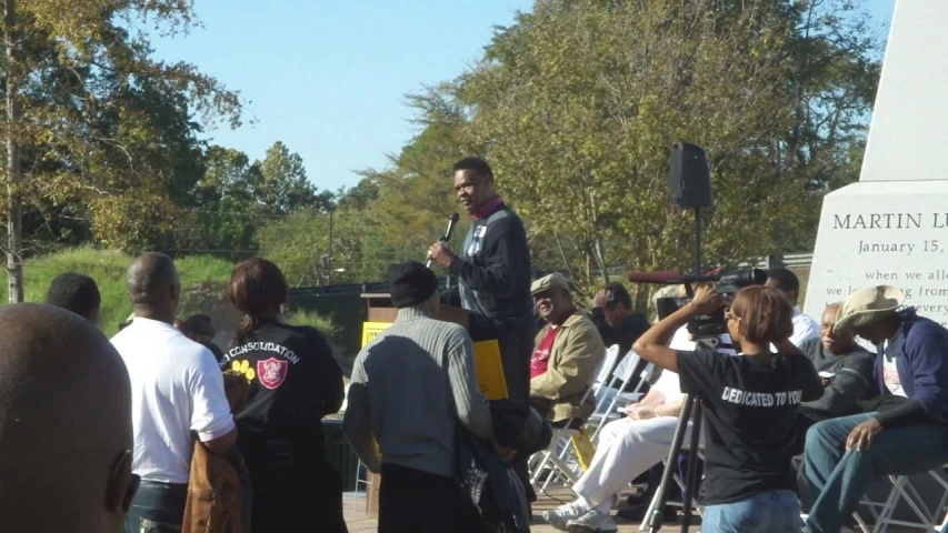 a crowd of people stand in the street as a man stands on stage and speaks