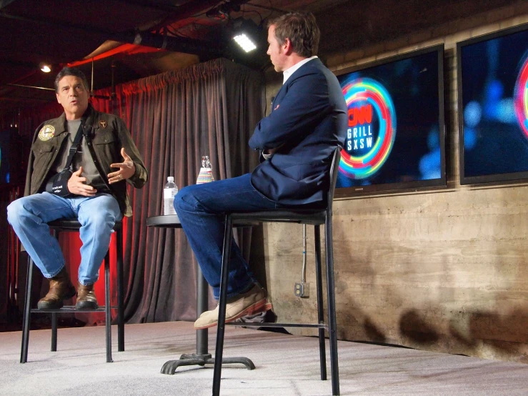 two men in suits sitting on a stage talking