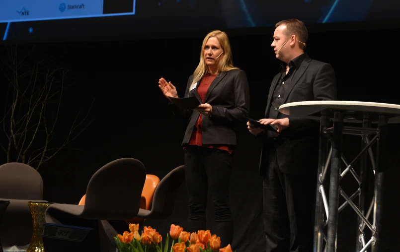 two people speaking on a stage next to a flower arrangement