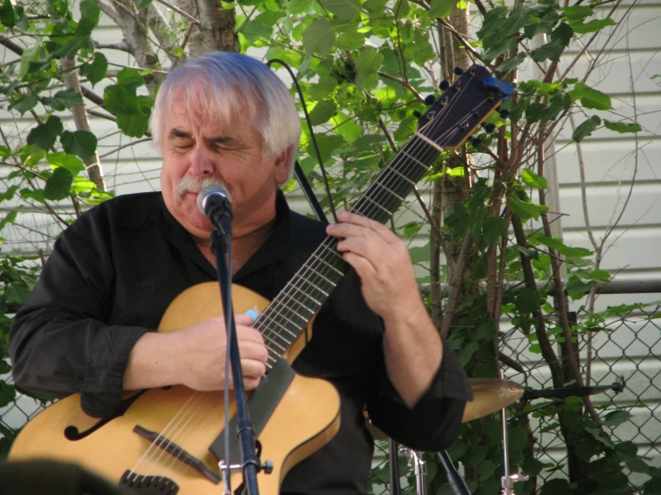 man with white hair holding an acoustic guitar while singing into microphone