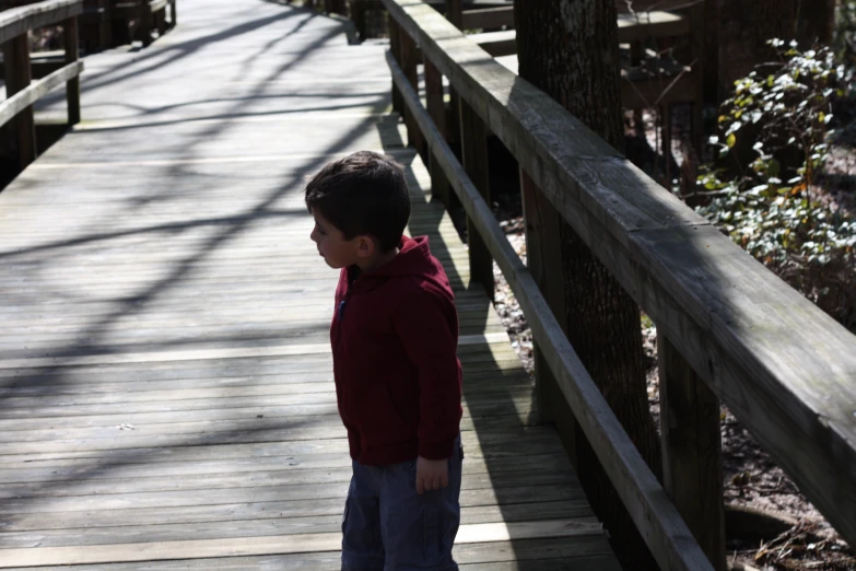 a boy in a red jacket looking at the ground on a wooden bridge