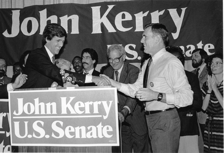 john kerry holding the us presidential glove while standing behind a sign