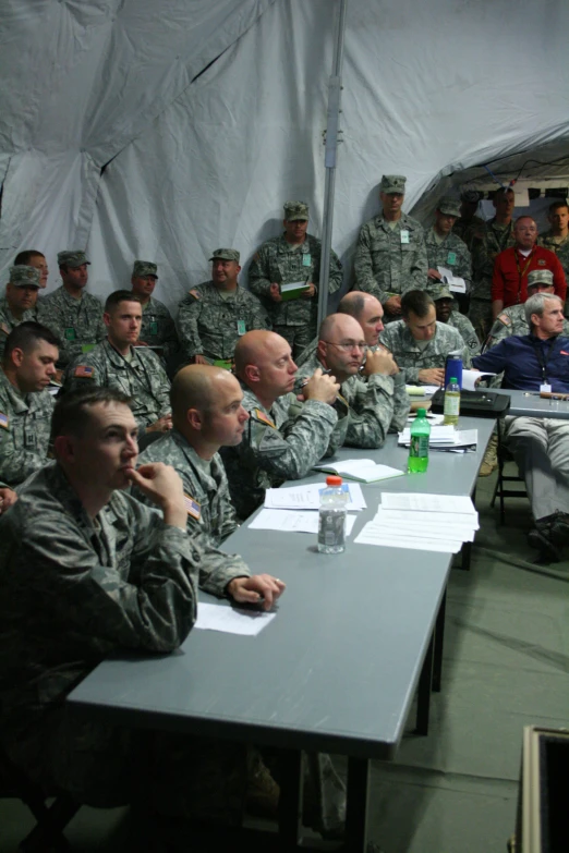 several men in camouflage uniforms are sitting at large tables in front of a wall with a man in red shirt on top