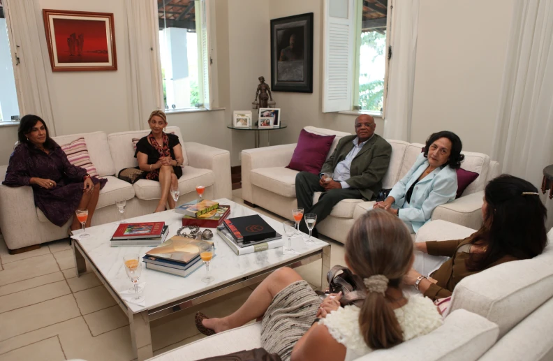 four women are seated in a room while two men sit down and a man standing up is talking