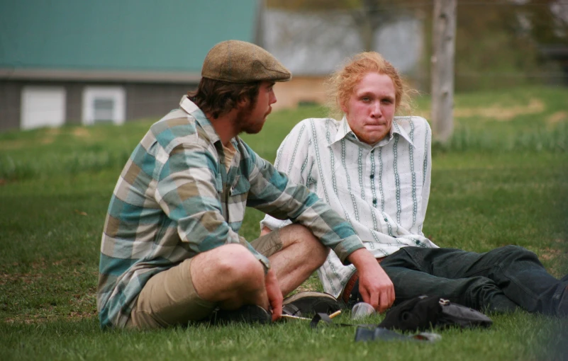 two people sit in the grass and one person holds a skateboard