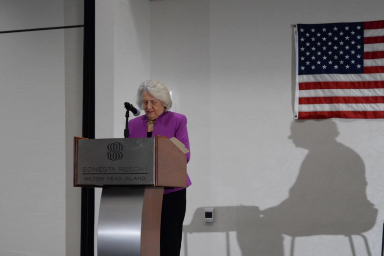 an elderly woman is giving a speech in front of a flag