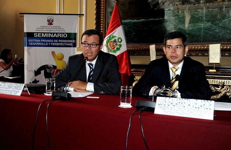 two men are sitting at a red table with papers