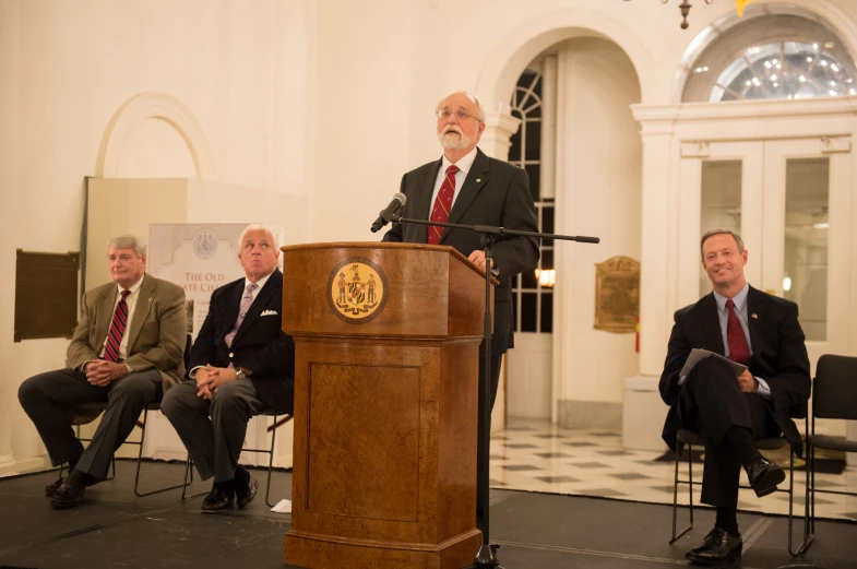 three men sit at a podium with microphones in front of them