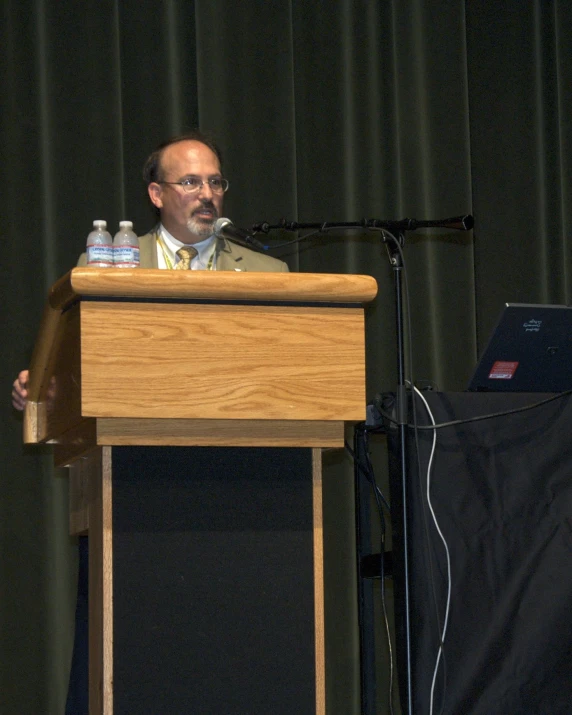 man standing at podium with microphone, with laptop open and holding bottle