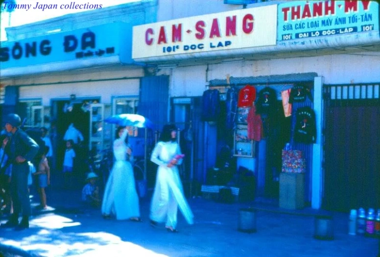 two women in white dresses holding umbrellas walk down a sidewalk